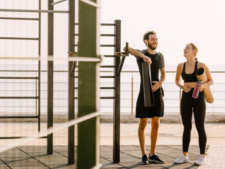 Ein Mann und eine Frau stehen im Freien neben einer Outdoor-Fitnessanlage. Beide halten jeweils eine Yogamatte in der Hand und lachen einander an.