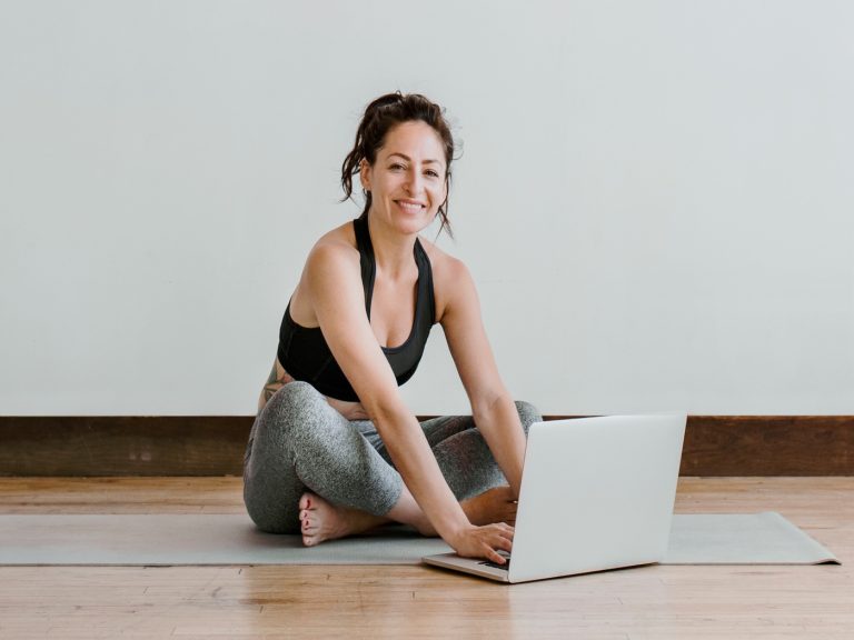 Frau sitz auf Yogamatte und arbeitet auf Laptop. Sie ist glücklich | yoga report